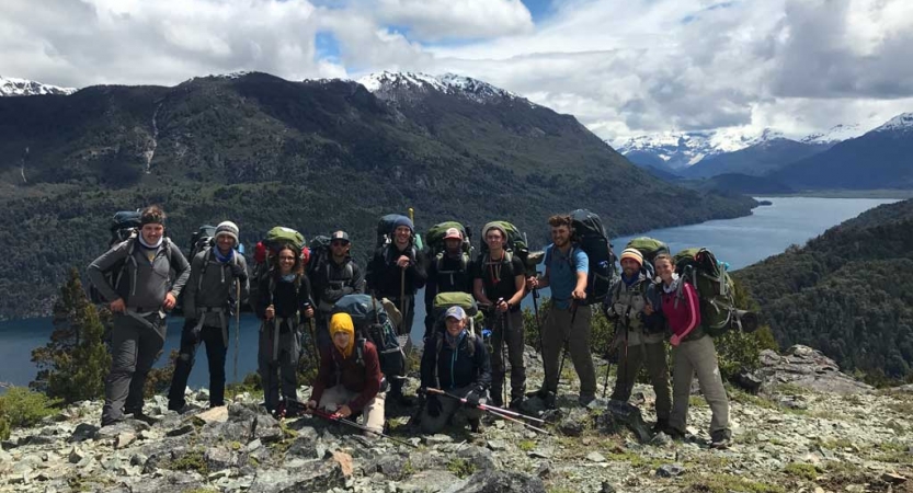 A group of people wearing backpacks pose for a group photo on an overlook. Behind them there are mountains and river. 